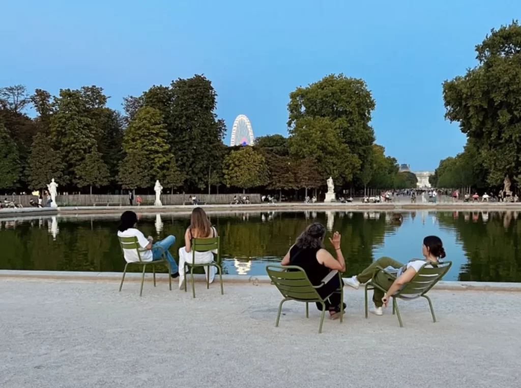 Visitantes sentados nas cadeiras verdes dos Jardins de Luxemburgo em Paris, com lago refletindo árvores e céu azul