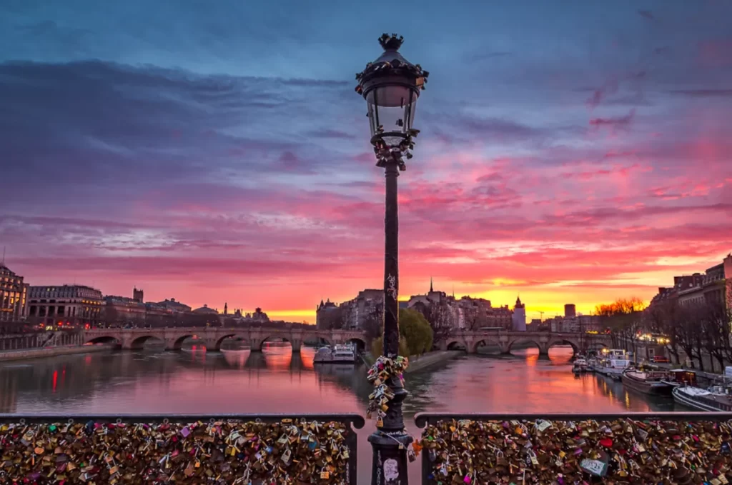 Ponte das Artes em Paris ao pôr do sol, com cadeados do amor, postes históricos e o rio Sena refletindo o céu colorido