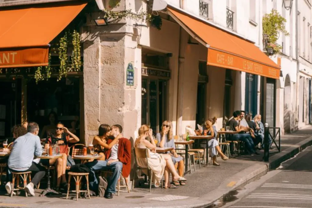 Um típico terraço de café parisiense em dia de sol, com mesas na calçada e o ritmo descontraído da cidade.