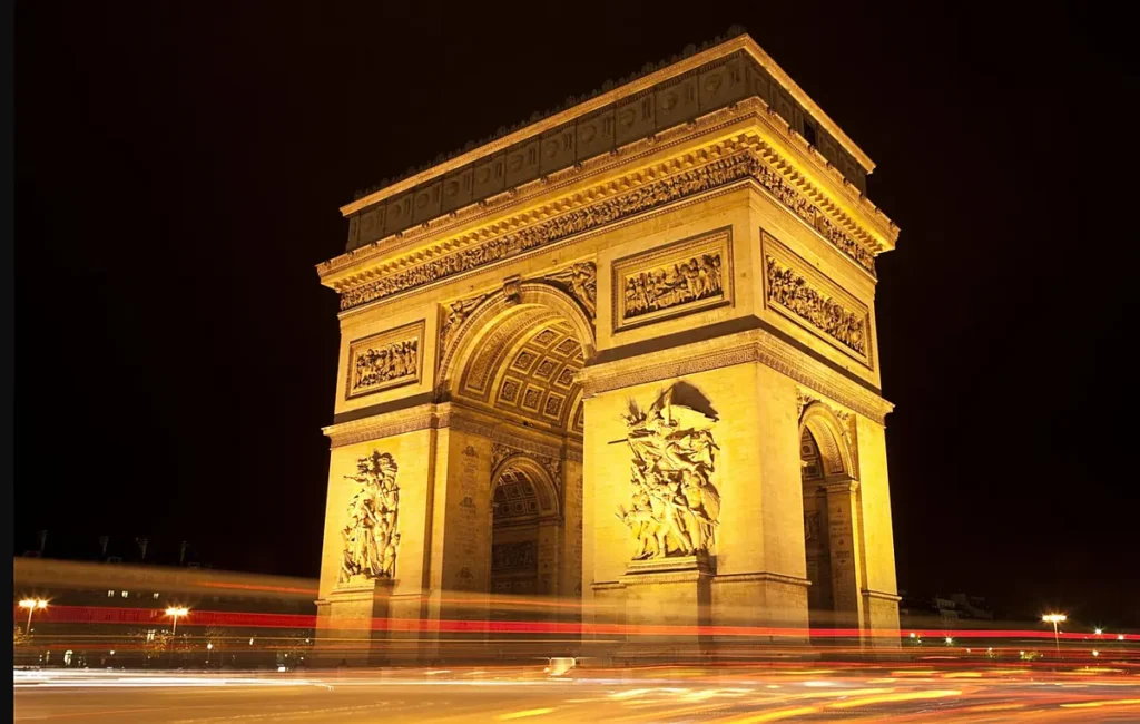 O Arco do Triunfo iluminado durante a noite em Paris, com suas esculturas monumentais e os rastros luminosos do trânsito na Place Charles de Gaulle.