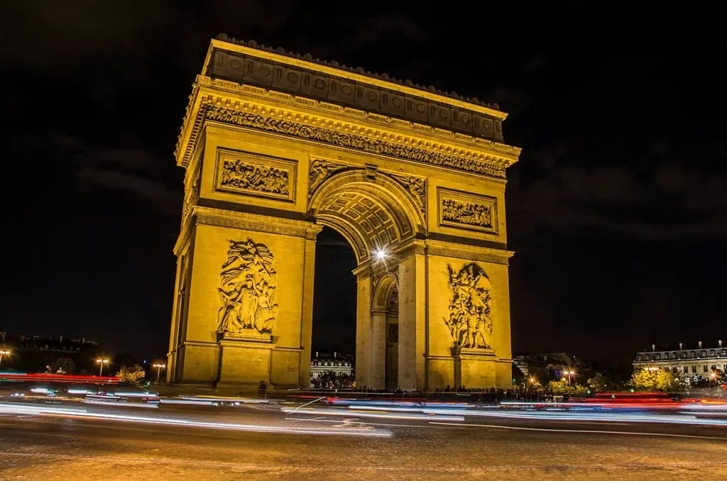 Vista lateral do Arco do Triunfo iluminado à noite em Paris, mostrando os relevos esculpidos, os detalhes arquitetônicos e os rastros de luz do trânsito. Copiar alt text