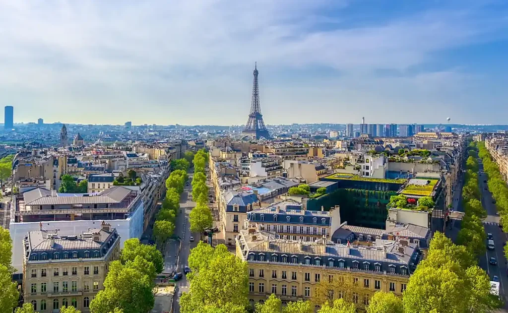 Vista aérea de Paris com a Torre Eiffel ao centro, telhados de edifícios históricos, avenidas arborizadas e o horizonte da capital francesa. Copiar alt text